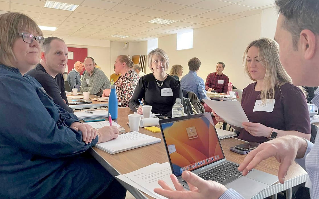 CDPS staff member discusses the Digital Service Standards for Wales with four others at a table at the CDPS Standards Roadshow. The room is filled with dozens of others discussing at their own tables and making notes.